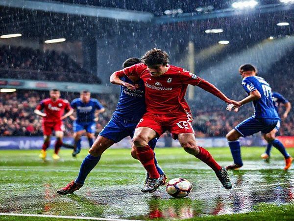 A high-definition image of a thrilling football match scene: two teams in vibrant kits, Stevenage in bright red and Cardiff in deep blue, clashing on a rain-soaked pitch under stadium lights. The atmosphere is electric, with passionate fans cheering from the stands. Focus on an intense moment where a Stevenage player tackles a Cardiff striker, showcasing the determination and skill of both teams. Capture the dynamic energy and emotion of the game, with droplets of rain reflecting the colorful lights.