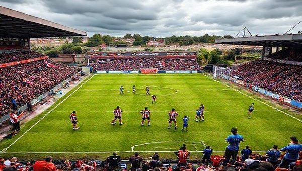 A vibrant football pitch at Whaddon Road, where the tension is palpable as Cheltenham Town's players clad in red and white prepare to face off against Buxton FC in their striking blue kit. The stadium is alive with passionate fans, waving flags and banners, both teams strategizing for a tactical battle. The scene captures an electrifying moment just before kickoff, with dramatic skies overhead, in high definition.