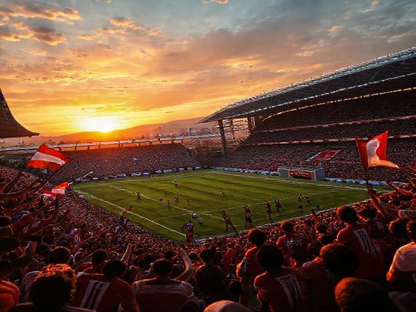 A vibrant football scene depicting a high-stakes match between FC Drita and AZ Alkmaar, filled with intense emotion. The stadium, packed with passionate fans waving flags, showcases Drita's "Warriors of Gjilan" in their striking kit, preparing for a counter-attack, while AZ’s players exhibit their intricate passing and attacking flair. The backdrop features a dramatic sunset over Gjilan, highlighting the tension and aspirations of both teams. Render this image in high definition.