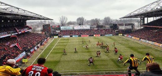 A dynamic football scene capturing the intense FA Cup match between Brackley Town and Burton Albion in December 2025. The stadium is buzzing with energy, showcasing passionate fans waving banners in the stands. On the field, Brackley, in their red and white kit, adopts a compact defensive formation, while Burton, in their yellow and black, showcases their intricate passing style. The clash of strategies is palpable, with focused players locked in a tactical duel, the cold breath of winter visible in the air. The atmosphere is electric, embodying the essence of grassroots football. High definition.
