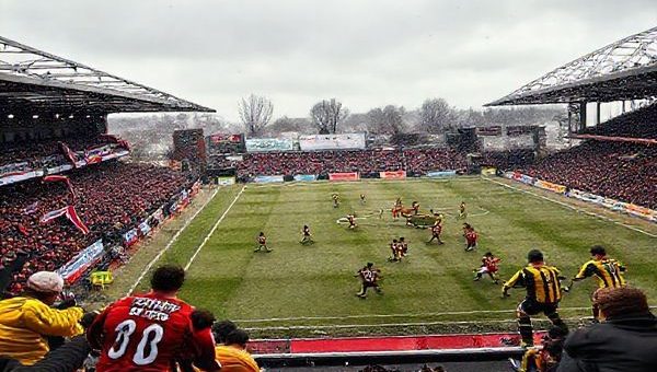 A dynamic football scene capturing the intense FA Cup match between Brackley Town and Burton Albion in December 2025. The stadium is buzzing with energy, showcasing passionate fans waving banners in the stands. On the field, Brackley, in their red and white kit, adopts a compact defensive formation, while Burton, in their yellow and black, showcases their intricate passing style. The clash of strategies is palpable, with focused players locked in a tactical duel, the cold breath of winter visible in the air. The atmosphere is electric, embodying the essence of grassroots football. High definition.