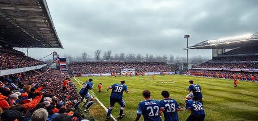 A dramatic football scene set in the Doonhamers' home ground under a crisp December sky, showcasing Queen of the South FC in navy blue jerseys, forming a disciplined defensive line, contrasted against the vibrant, energetic Cove Rangers in bright orange, launching a fast-paced attack. The stadium is packed with passionate fans, flags waving, as anticipation fills the air. Include dynamic movements, a classic football atmosphere, and the intensity of the clash, all in high definition.