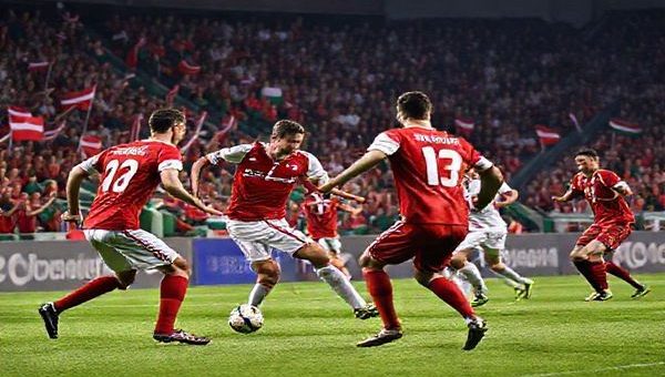 A tense football match scene showcasing Denmark versus Belarus in a World Cup qualifier, with Danish players in red and white jerseys executing a precise attack, while Belarus players in red and green defend vigorously. The stadium is packed with enthusiastic fans waving flags, creating an electric atmosphere. Capture the essence of tactical chess on the field, highlighting Denmark's efficiency and Belarus's gritty counter-attacks. Make the image in high definition.