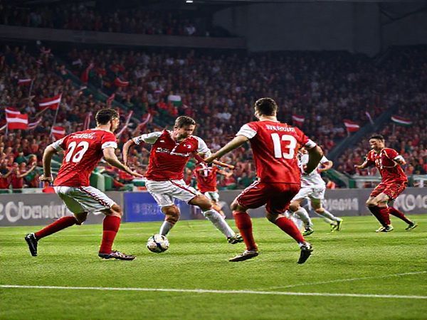 A tense football match scene showcasing Denmark versus Belarus in a World Cup qualifier, with Danish players in red and white jerseys executing a precise attack, while Belarus players in red and green defend vigorously. The stadium is packed with enthusiastic fans waving flags, creating an electric atmosphere. Capture the essence of tactical chess on the field, highlighting Denmark's efficiency and Belarus's gritty counter-attacks. Make the image in high definition.