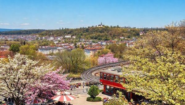 A picturesque view of Wiesbaden in spring, featuring blooming flowers and budding trees in vibrant colors. In the foreground, locals and tourists enjoy sunny outdoor cafés. The historic Nerobergbahn rail climbs a nearby hill, offering stunning panoramic views of the charming cityscape with neoclassical architecture and lush parks. Spring festivities fill the air, with artists showcasing their work and joyous events taking place in the background. A gentle breeze stirs the leaves, and the atmosphere is alive with the promise of renewal and cultural richness.