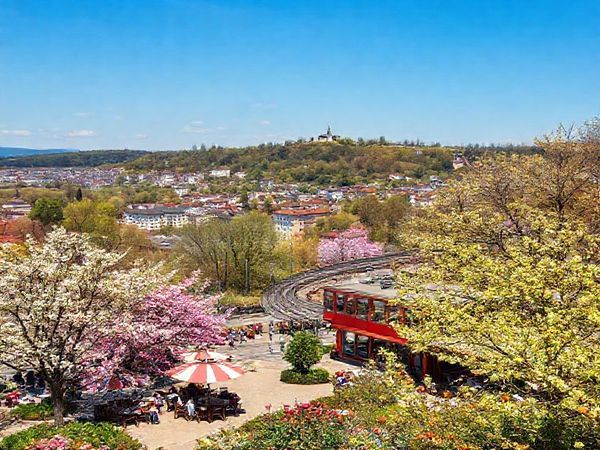 A picturesque view of Wiesbaden in spring, featuring blooming flowers and budding trees in vibrant colors. In the foreground, locals and tourists enjoy sunny outdoor cafés. The historic Nerobergbahn rail climbs a nearby hill, offering stunning panoramic views of the charming cityscape with neoclassical architecture and lush parks. Spring festivities fill the air, with artists showcasing their work and joyous events taking place in the background. A gentle breeze stirs the leaves, and the atmosphere is alive with the promise of renewal and cultural richness.