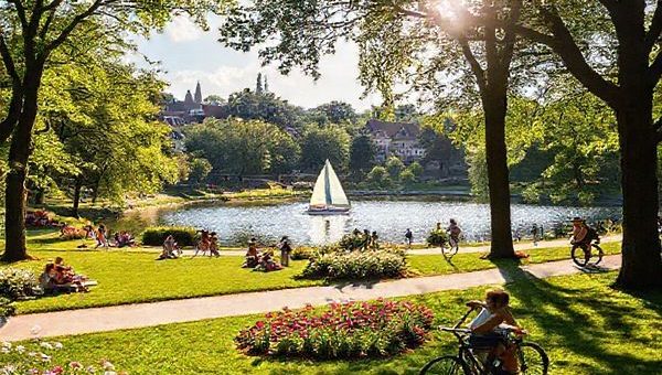 A vibrant scene of Krefeld, Germany in summer—a picturesque park filled with lush greenery and blossoming flowers, families enjoying picnics under tall trees, and cyclists navigating well-marked bike paths. The background features the historic architecture of the city, with hints of its textile heritage, while children play by the sparkling Elfrather See, where a sailing boat glides across the water. A warm sun casts gentle rays, creating a lively, welcoming atmosphere, symbolizing the delightful blend of nature, culture, and community in this charming city.