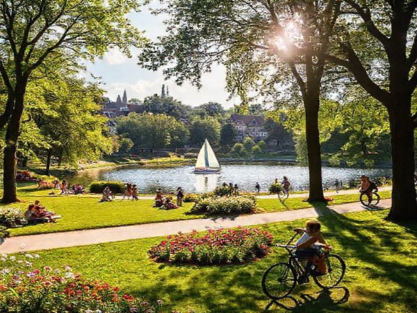 A vibrant scene of Krefeld, Germany in summer—a picturesque park filled with lush greenery and blossoming flowers, families enjoying picnics under tall trees, and cyclists navigating well-marked bike paths. The background features the historic architecture of the city, with hints of its textile heritage, while children play by the sparkling Elfrather See, where a sailing boat glides across the water. A warm sun casts gentle rays, creating a lively, welcoming atmosphere, symbolizing the delightful blend of nature, culture, and community in this charming city.