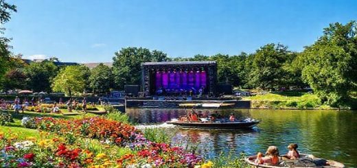 A high-definition image of Dortmund in summer, showcasing the vibrant Westfalenpark filled with colorful flowers, locals and tourists enjoying picnics, and a boat sailing leisurely along the Emscher River. In the background, there’s a lively outdoor concert at Signal Iduna Park, surrounded by lush greenery and a clear blue sky, capturing the dynamic energy of summer in the city.