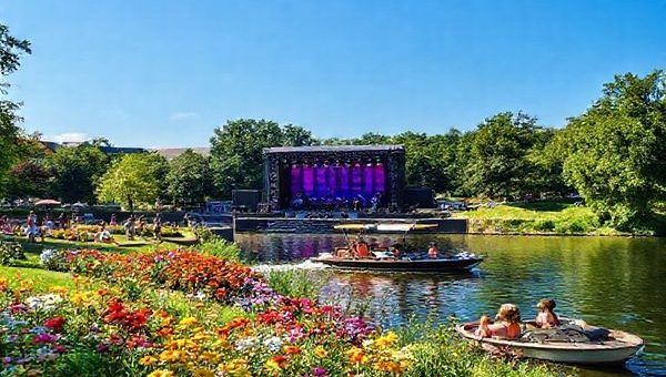 A high-definition image of Dortmund in summer, showcasing the vibrant Westfalenpark filled with colorful flowers, locals and tourists enjoying picnics, and a boat sailing leisurely along the Emscher River. In the background, there’s a lively outdoor concert at Signal Iduna Park, surrounded by lush greenery and a clear blue sky, capturing the dynamic energy of summer in the city.