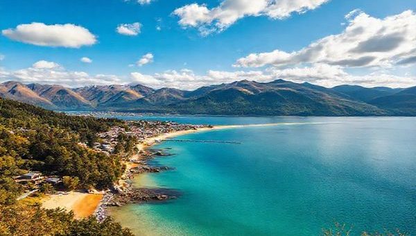 Create a high-definition image of the stunning landscapes of Nelson District, New Zealand, showcasing the vibrant colors of summer with golden beaches, lush forests, and serene mountains. Include a lively local market bustling with crafts and produce, and the crystal-clear waters of Abel Tasman National Park. Capture the essence of outdoor adventures and a relaxed Kiwi lifestyle, under a brilliant blue sky filled with fluffy clouds.