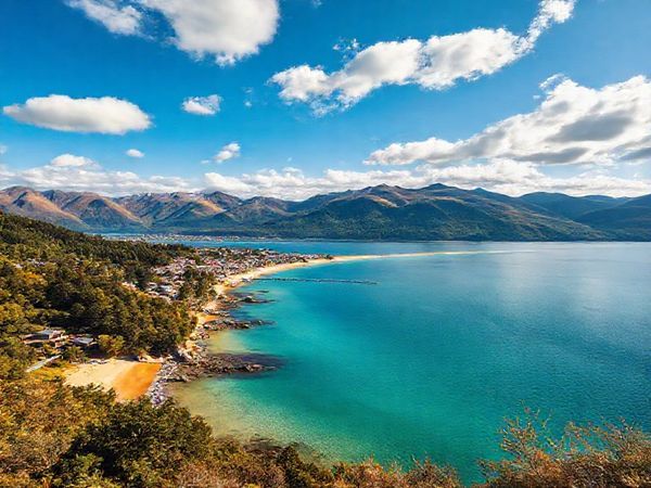 Create a high-definition image of the stunning landscapes of Nelson District, New Zealand, showcasing the vibrant colors of summer with golden beaches, lush forests, and serene mountains. Include a lively local market bustling with crafts and produce, and the crystal-clear waters of Abel Tasman National Park. Capture the essence of outdoor adventures and a relaxed Kiwi lifestyle, under a brilliant blue sky filled with fluffy clouds.