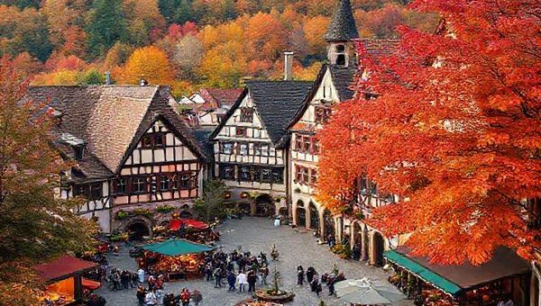Create a high-definition image of Villingen-Schwenningen, Germany, showcasing the enchanting medieval architecture blanketed in colorful autumn leaves of deep red, orange, and gold. Include a vibrant town square bustling with visitors enjoying food stalls, wood fires, and festive decorations. The backdrop should feature the lush Black Forest, with trails meandering through the picturesque landscape. Capture the essence of fall in this magical destination, highlighting both the natural beauty and cultural vibrancy.