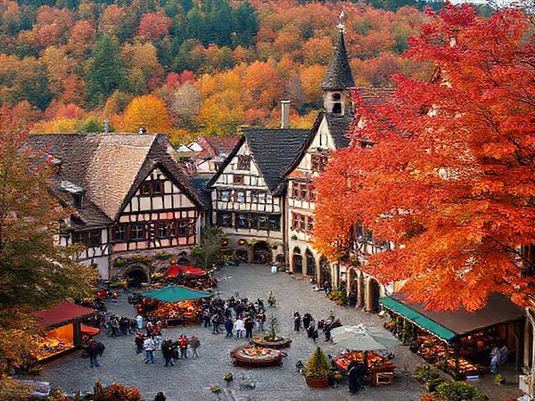 Create a high-definition image of Villingen-Schwenningen, Germany, showcasing the enchanting medieval architecture blanketed in colorful autumn leaves of deep red, orange, and gold. Include a vibrant town square bustling with visitors enjoying food stalls, wood fires, and festive decorations. The backdrop should feature the lush Black Forest, with trails meandering through the picturesque landscape. Capture the essence of fall in this magical destination, highlighting both the natural beauty and cultural vibrancy.
