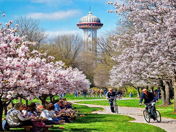 "A vibrant scene of Oberhausen in spring, showcasing cherry blossoms and magnolias in full bloom, with locals enjoying picnics in a park. In the background, the Gasometer stands tall, adorned with colorful decorations. Cyclists are seen along the Emscher Park Bicycle Trail, surrounded by lush landscapes. Capture the energy and beauty of this lively city in high definition."
