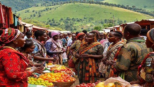 A vibrant market scene in Rwanda, showcasing locals engaging in conversation while surrounded by colorful stalls of fruits, textiles, and traditional crafts. In the foreground, a diverse group of learners, both local and international, practice Kinyarwanda with native speakers, embodying the spirit of cultural exchange. Lush green hills and the iconic Rwandan landscape create a stunning backdrop, emphasizing the beauty of immersing oneself in the culture. High definition.