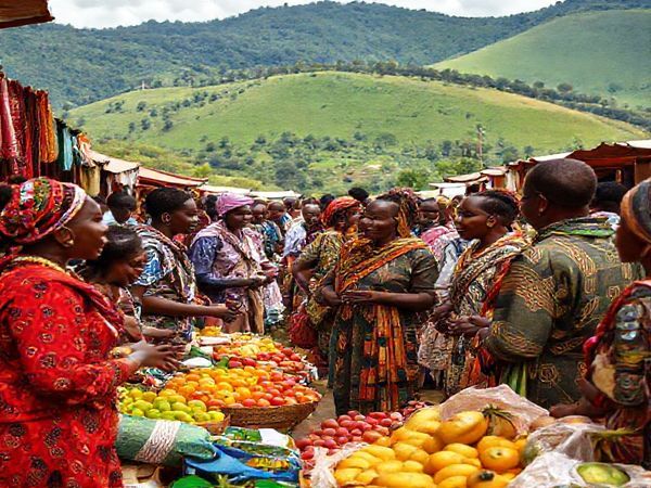 A vibrant market scene in Rwanda, showcasing locals engaging in conversation while surrounded by colorful stalls of fruits, textiles, and traditional crafts. In the foreground, a diverse group of learners, both local and international, practice Kinyarwanda with native speakers, embodying the spirit of cultural exchange. Lush green hills and the iconic Rwandan landscape create a stunning backdrop, emphasizing the beauty of immersing oneself in the culture. High definition.