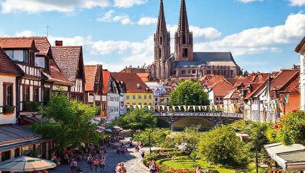 A high-definition depiction of Erfurt, Germany, showcasing the vibrant medieval architecture and cobblestone streets. Illustrate the city during summer, with bustling outdoor cafes, lush green parks in full bloom, and colorful festival banners, while capturing the iconic spires of Erfurt Cathedral and the charming Kramer Bridge in the background. Infuse the scene with warm sunlight and joyful tourists enjoying the lively atmosphere.