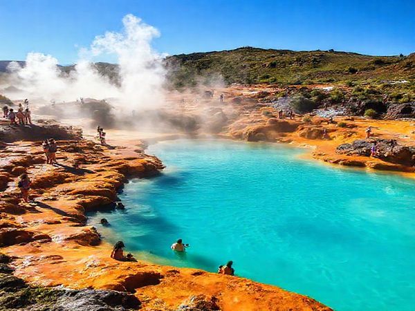 Create a high-definition image of Champagne Pool in New Zealand, capturing the vibrant hues of its steaming turquoise waters surrounded by deep oranges, vivid yellows, and greens of the mineral deposits. Envision a lively summer scene with tourists joyfully exploring the area, set against a backdrop of a clear blue sky and warm sunlight. Incorporate hints of geothermal steam rising from the pool, alongside lush flora. The atmosphere should convey energy, excitement, and a sense of adventure amidst this geothermal wonderland.