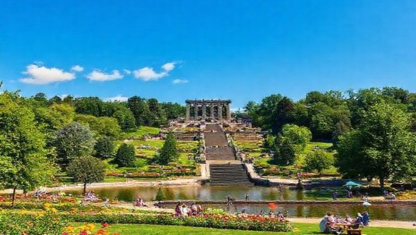 Create a high definition image of Kassel, Germany, showcasing the enchanting Bergpark Wilhelmshöhe in summer, complete with lush gardens, the grand Hercules monument, and people enjoying picnics by the Fulda River. The scene should be vibrant and alive, featuring colorful flowers and a clear blue sky, capturing the energy of festivals and outdoor events in this culturally rich city.