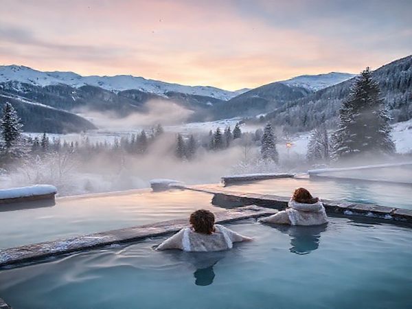 A serene view of Hanmer Springs Thermal Pools during winter, showcasing snow-capped mountains surrounding steaming thermal waters. Include a few wispy snowflakes falling, with a couple of visitors relaxing in the warm pools, wrapped in cozy robes. The landscape is tranquil and magical with frosted trees and a hint of evening dusk light. High definition.