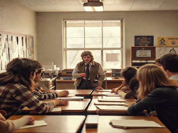 Create a high-definition image of a nostalgic 1970s classroom scene, capturing the essence of a gruff teacher, Mr. Harrington, with a vintage aesthetic, surrounded by students engaged in lively discussion. In the corner, a strong yet nurturing female figure, Mary, watches over them, embodying both resilience and empathy. The walls are adorned with retro posters, and sunlight filters through dusty windows, casting warm tones that evoke a sense of longing and reflection on gender dynamics and societal expectations.
