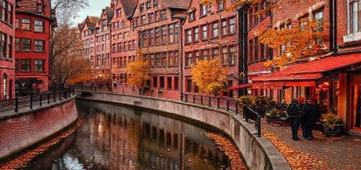 A high-definition image of Speicherstadt in autumn, showcasing a picturesque canal lined with historic red-brick warehouses adorned in vibrant hues of amber and crimson. The scene captures fallen leaves drifting along the cobbled streets, with a couple leisurely walking and a cozy café in the background, emphasizing the enchanting allure of fall in this UNESCO World Heritage site.
