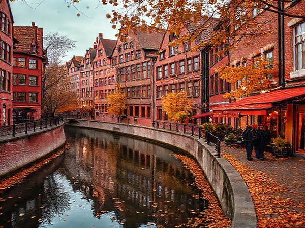 A high-definition image of Speicherstadt in autumn, showcasing a picturesque canal lined with historic red-brick warehouses adorned in vibrant hues of amber and crimson. The scene captures fallen leaves drifting along the cobbled streets, with a couple leisurely walking and a cozy café in the background, emphasizing the enchanting allure of fall in this UNESCO World Heritage site.