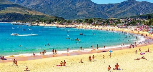 A vibrant summer scene in Waipu, New Zealand, showcasing golden sandy beaches and clear blue waters at Bream Bay. Include lively beachgoers enjoying water sports, with the mountains in the background and colorful flags from the Waipu Christmas Parade fluttering in the gentle breeze. Capture the essence of a sunny day with bright skies and a bustling atmosphere, reflecting the town's Scottish heritage. Create in high definition.