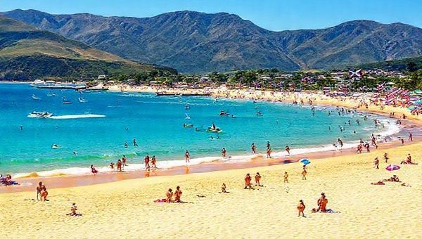 A vibrant summer scene in Waipu, New Zealand, showcasing golden sandy beaches and clear blue waters at Bream Bay. Include lively beachgoers enjoying water sports, with the mountains in the background and colorful flags from the Waipu Christmas Parade fluttering in the gentle breeze. Capture the essence of a sunny day with bright skies and a bustling atmosphere, reflecting the town's Scottish heritage. Create in high definition.