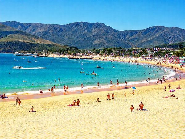 A vibrant summer scene in Waipu, New Zealand, showcasing golden sandy beaches and clear blue waters at Bream Bay. Include lively beachgoers enjoying water sports, with the mountains in the background and colorful flags from the Waipu Christmas Parade fluttering in the gentle breeze. Capture the essence of a sunny day with bright skies and a bustling atmosphere, reflecting the town's Scottish heritage. Create in high definition.