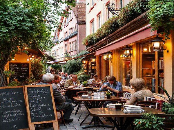 Create a high definition image of a cozy Luxembourgish café scene, bustling with locals engaging in conversation, surrounded by beautiful traditional architecture and lush greenery. Include elements like language learning books, a chalkboard with Luxembourgish phrases, and warm lighting reflecting a welcoming atmosphere. Make the setting vibrant and inviting, emphasizing the cultural richness of Luxembourg.