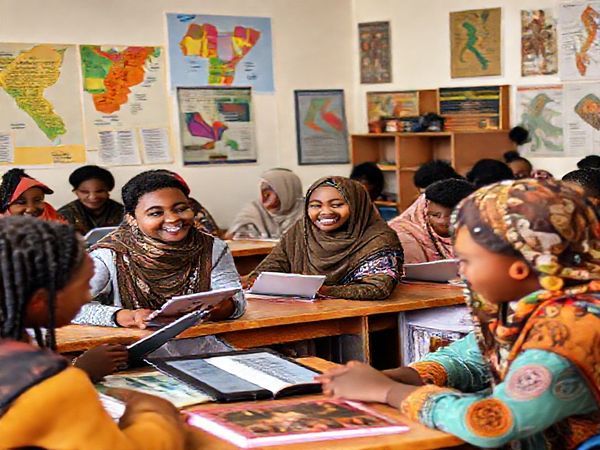 Create a high definition image of a vibrant classroom setting with diverse students engaged in learning Somali. The walls are adorned with colorful maps of Somalia and posters showcasing Somali poetry and traditional art. In the foreground, a cheerful teacher assists a student with pronunciation, while other students practice speaking with each other. The atmosphere is lively, filled with books, language learning apps on tablets, and cultural items like traditional Somali artifacts, depicting the richness of Somali heritage and education.