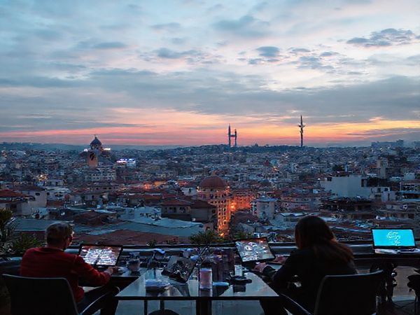 A vibrant cityscape of Türkiye's bustling tech hubs, where traditional architecture meets modern digital innovation. Visualize an array of entrepreneurs working on laptops in co-working spaces, digital screens displaying social media logos, and diverse professionals engaged in discussions. Incorporate glimpses of rich Turkish culture alongside futuristic technology, illustrating the fusion of old and new. The sky is a blend of twilight hues, symbolizing a new era. Create this scene in high definition.