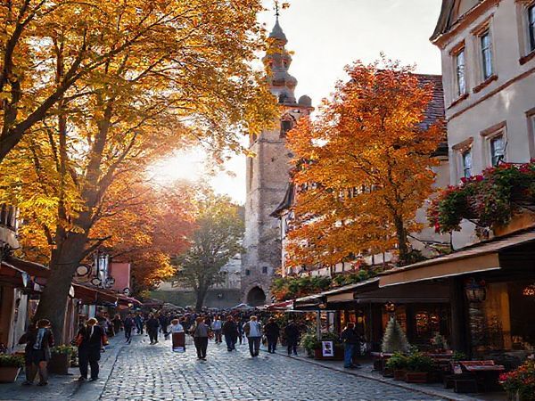 Create a high-definition image of Augsburg during the enchanting autumn season, featuring a vibrant tapestry of colorful leaves in the parks, historical Renaissance architecture, and a charming street scene bustling with people enjoying local Oktoberfest celebrations. Soft, golden sunlight filters through the trees, casting a warm glow over the cobblestone streets, with cozy cafes and art galleries inviting exploration in the background. Include elements like traditional Bavarian decor and the iconic Perlach Tower to capture the city's rich cultural heritage amidst the fall atmosphere.