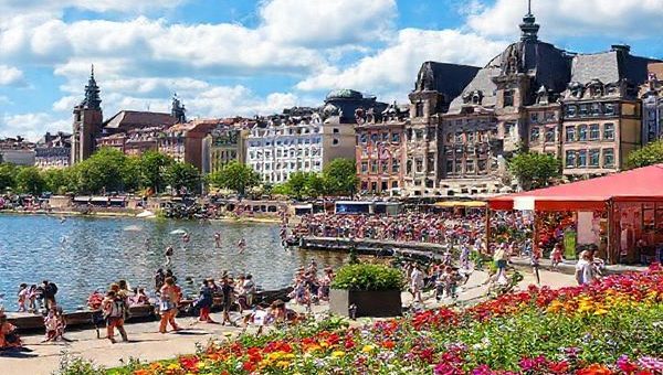 Create a high definition image of a vibrant scene in Hamburg, depicting the bustling summer atmosphere with people enjoying outdoor activities on Alster Lake, colorful flowers blooming in Planten un Blomen Park, and iconic historic architecture in the background. Include festive elements such as music and art displays celebrating the Altonale festival, under a bright blue sky filled with fluffy white clouds, capturing the essence of joy and vibrancy in this captivating port city.