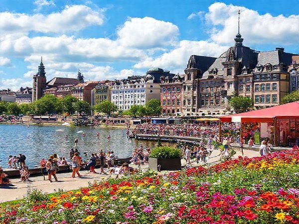 Create a high definition image of a vibrant scene in Hamburg, depicting the bustling summer atmosphere with people enjoying outdoor activities on Alster Lake, colorful flowers blooming in Planten un Blomen Park, and iconic historic architecture in the background. Include festive elements such as music and art displays celebrating the Altonale festival, under a bright blue sky filled with fluffy white clouds, capturing the essence of joy and vibrancy in this captivating port city.