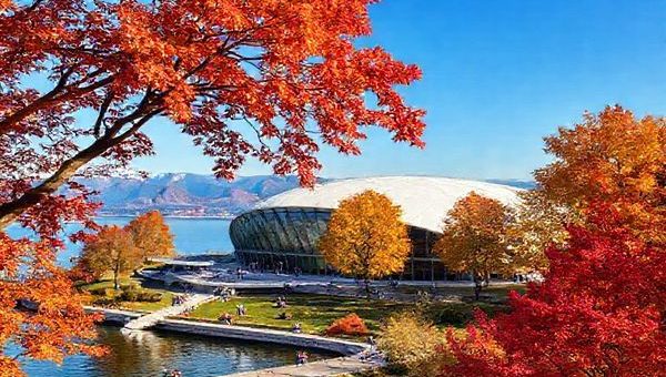 A stunning scene depicting the Zeppelin Museum nestled on the shores of Lake Constance during a vibrant autumn day. Show the museum's architecture surrounded by trees in shades of red, orange, and gold, with visitors leisurely exploring the exhibits and enjoying the beautiful outdoors. Include a glimpse of the lake reflecting the colorful foliage and distant mountains under a clear blue sky. Make the image in high definition.