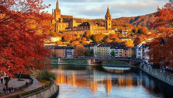 "An enchanting view of Heidelberg, Germany, showcasing the stunning Heidelberg Castle and the Neckar River, surrounded by vibrant autumn foliage in hues of orange, red, and gold. Include charming medieval architecture and people enjoying a leisurely stroll along the riverbank. Incorporate a soft golden light of early evening, enhancing the magical atmosphere. Render this scene in high definition."