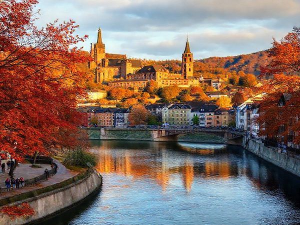 "An enchanting view of Heidelberg, Germany, showcasing the stunning Heidelberg Castle and the Neckar River, surrounded by vibrant autumn foliage in hues of orange, red, and gold. Include charming medieval architecture and people enjoying a leisurely stroll along the riverbank. Incorporate a soft golden light of early evening, enhancing the magical atmosphere. Render this scene in high definition."
