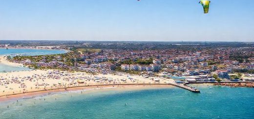 "A panoramic view of the Baltic Coast in summer, showcasing vibrant beach scenes filled with sunbathers and families enjoying the golden sands, lively local markets, and a backdrop of charming seaside towns like Lübeck. The azure sea sparkles under a clear blue sky, while colorful kites soar above adventurous kite surfers. High definition."