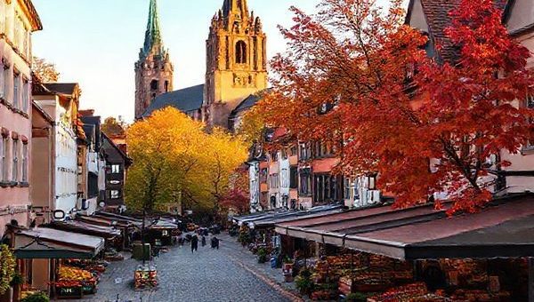 A picturesque view of Wittenberg, Germany, during autumn, showcasing cobblestone streets lined with medieval architecture adorned in vibrant shades of gold and crimson leaves. The scene captures local markets bustling with fresh produce, while an iconic church stands in the background, bathed in the soft glow of early evening light. High-definition.