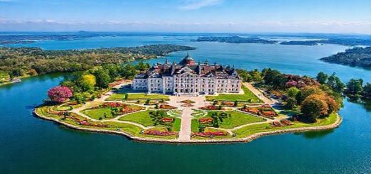 A stunning aerial view of Herrenchiemsee Island in Bavaria, showcasing the opulent Herrenchiemsee Palace surrounded by vibrant summer gardens in full bloom, the shimmering Chiemsee lake reflecting the clear blue sky, and tourists enjoying leisurely strolls, all depicted in high definition with vivid colors and intricate details.