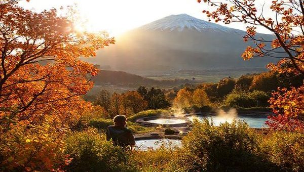 A breathtaking high-definition landscape of Kawerau District in New Zealand, showcasing the vibrant colors of autumn with golden and amber leaves on trees, the majestic Tarawera Mountain in the background, and the serene hot springs nestled among lush greenery. Capture a tranquil scene of a visitor enjoying nature, framed by the inviting warm light of the late afternoon sun, with local wildlife subtly included in the foreground.