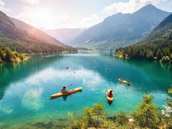 A breathtaking panoramic view of Eibsee Lake nestled in the Bavarian Alps, showcasing vibrant summer colors with families enjoying water activities such as kayaking and paddleboarding. The crystal-clear waters reflect the lush greenery and rugged mountain peaks, while sunlit trails meander through the surrounding forest, creating a lively and festive atmosphere. High definition.