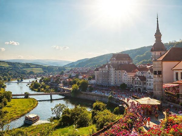 A breathtaking high-definition scene showcasing Landkreis Regensburg in summer, with vibrant landscapes bathed in golden sunlight. Capture the lush green countryside, the serene Danube River with a leisurely boat, bustling historical towns celebrating local festivals, and joyful parades filled with colorful decorations. Include architectural marvels like Walhalla under the summer sun, surrounded by blooming flowers and people enjoying outdoor activities.